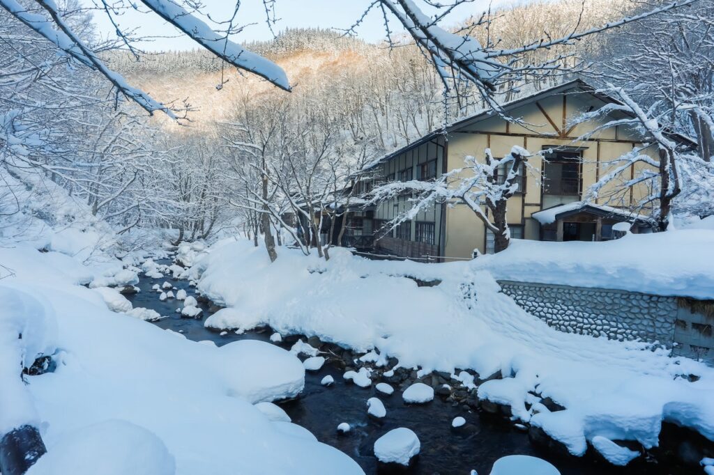 冬の雪景色に包まれた温泉旅館の風景。木々や小川が雪で覆われ、静寂で美しい冬の自然に囲まれた情緒ある建物が川沿いに建っています