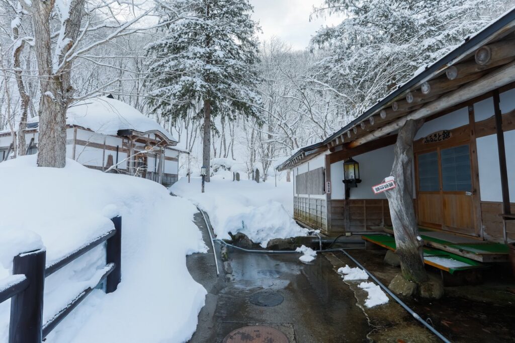 冬の雪景色に包まれた温泉旅館の風景。木々や小川が雪で覆われ、静寂で美しい冬の自然に囲まれた情緒ある建物が川沿いに建っています。さっきと画像と角度違い